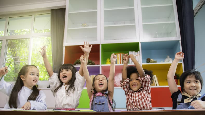 Group of multi-ethnic little kids raising their hands in happiness in a classroom